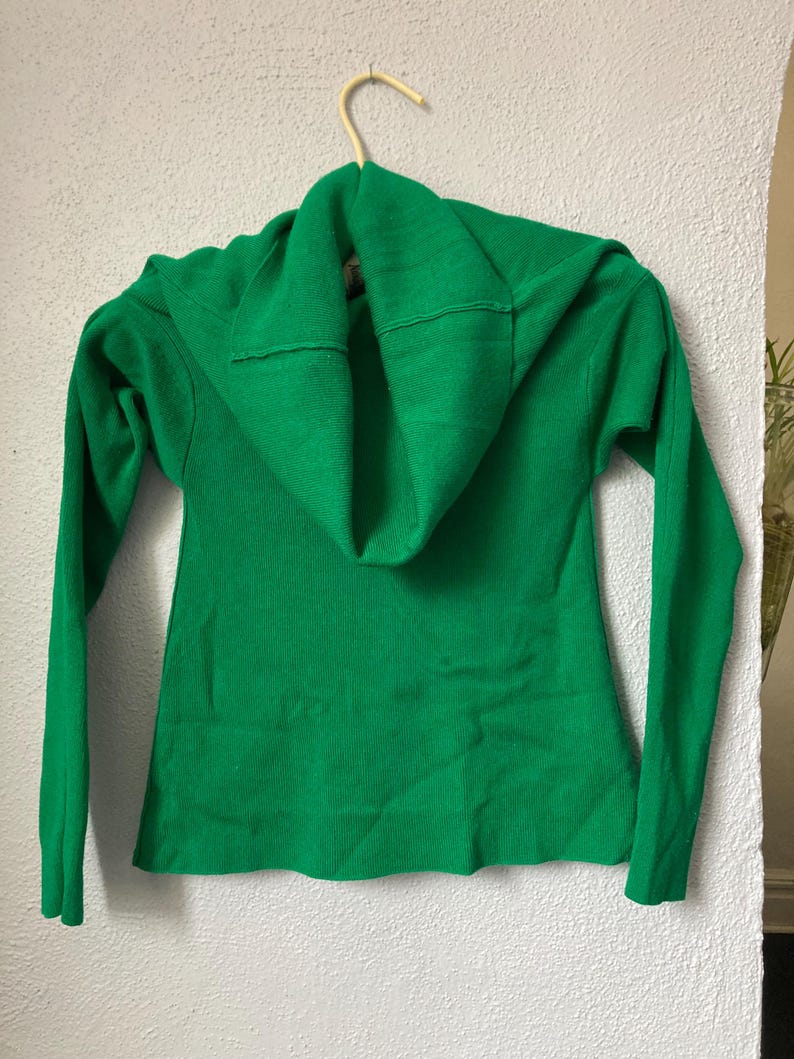 May include: A vibrant green sweater with a cowl neck, hanging on a wooden hanger against a white wall. The long-sleeved sweater features a ribbed texture and a folded cowl neckline. The garment is made of a soft, knitted material.