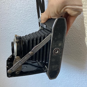 May include: A vintage black folding camera with a black strap. The camera has a bellows design and silver-colored metal accents. The camera is held by a person.