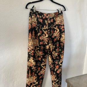 May include: Black pants with a colorful floral and architectural pattern. The pants are hanging on a black hanger against a white wall. The pattern includes red, beige, and gold elements.
