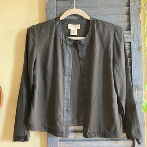 May include: A black long-sleeve jacket with a round collar and button closure. The jacket has a satin trim along the front and cuffs. The garment is hanging on a white hanger against a blue shutter background.