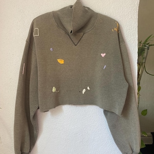 May include: An olive green cropped sweatshirt with a mock neck collar and long sleeves. The garment features small, scattered hand-embroidered details in various colors including yellow, pink, white, and purple, with a raw, unfinished hemline.