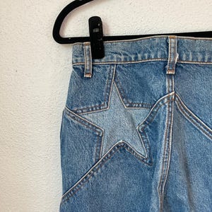 May include: Blue denim jeans featuring a large, lighter-toned star-shaped patch design on the hip area. The jeans have a high-waisted cut with visible belt loops and thick, contrasting orange-brown stitching along the seams and star pattern.