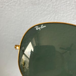 Op de afbeelding: Close-up van een Ray-Ban zonnebril met gouden montuur en groene glazen. Het Ray-Ban logo is zichtbaar op de lens. De zonnebril heeft een klassieke aviator stijl.