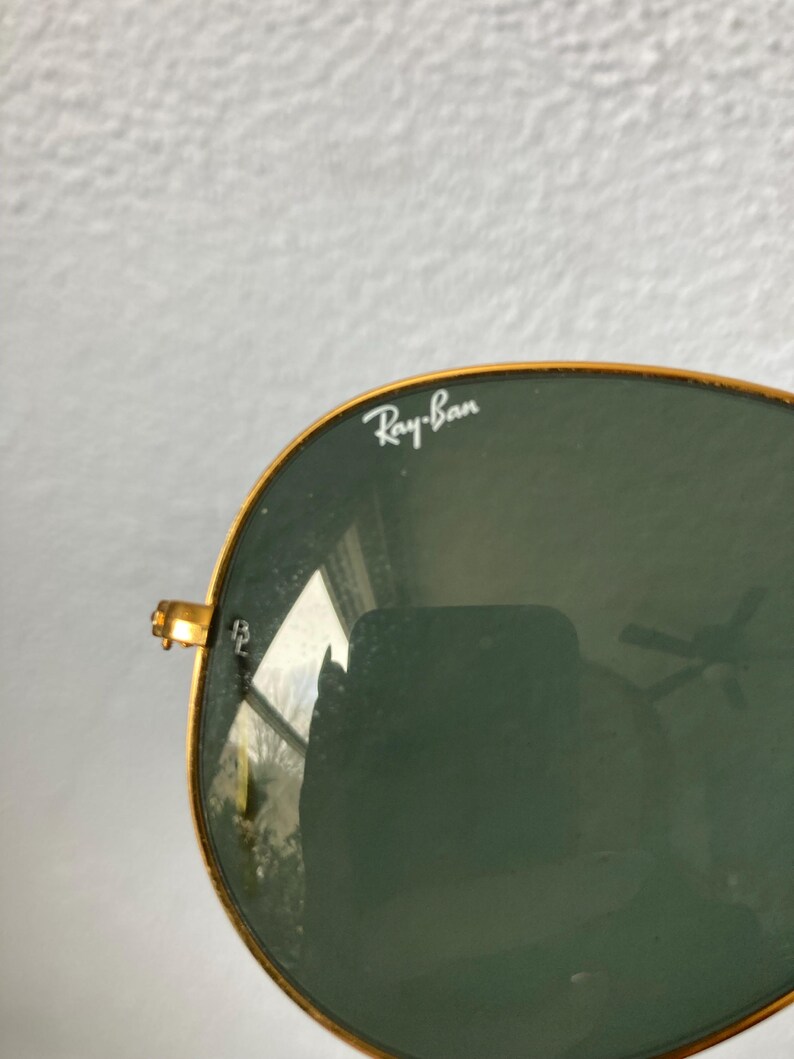 Op de afbeelding: Close-up van een Ray-Ban zonnebril met gouden montuur en groene glazen. Het Ray-Ban logo is zichtbaar op de lens. De zonnebril heeft een klassieke aviator stijl.
