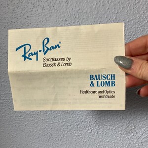 Op de afbeelding: Een gevouwen papieren advertentie voor Ray-Ban zonnebrillen van Bausch & Lomb. Het Ray-Ban-logo is blauw, met de tekst "Sunglasses by Bausch & Lomb" eronder. Het onderste gedeelte toont het Bausch & Lomb-logo en tekst.