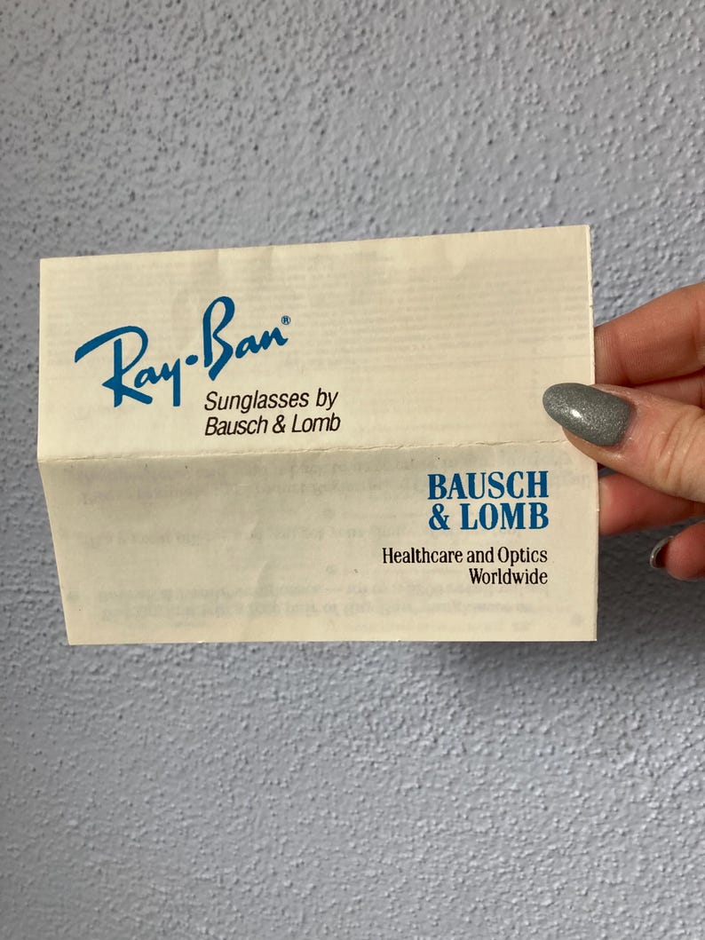 Op de afbeelding: Een gevouwen papieren advertentie voor Ray-Ban zonnebrillen van Bausch & Lomb. Het Ray-Ban-logo is blauw, met de tekst "Sunglasses by Bausch & Lomb" eronder. Het onderste gedeelte toont het Bausch & Lomb-logo en tekst.