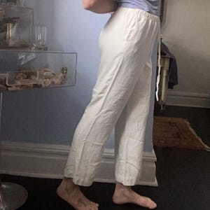 May include: Off-white, loose-fitting pants with a drawstring waist. The pants are made of a lightweight fabric and have a relaxed fit. The pants are shown from the side, with the person wearing them partially visible.