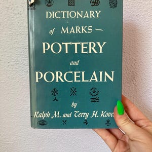 May include: A teal hardcover book titled Dictionary of Marks - Pottery and Porcelain by Ralph M. and Terry H. Kovel. The cover features various black printed pottery marks and symbols, with a small tear on the top left corner of the dust jacket.