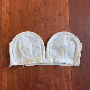 May include: Ivory-colored vintage bra with a lace overlay. The bra features a satin ribbon and bow detail at the center. The bra is laid flat on a wooden surface.