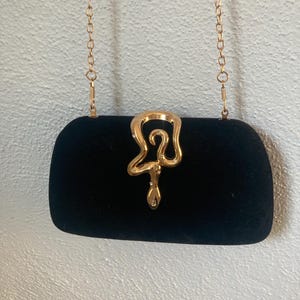 May include: A black velvet clutch bag featuring a gold-toned metal snake-shaped clasp on the front. The bag includes a thin gold-toned metal chain strap attached to the top, allowing it to be carried or worn over the shoulder as a handbag.