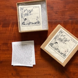 May include: Two small, square, cardboard boxes with a printed label featuring a black and white illustration and the text "ex libris." One box is open, revealing the label inside. A small piece of paper with text is also present.