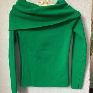 May include: A vibrant green off-the-shoulder jumper with long sleeves. The garment is suspended on a beige hanger against a light grey wall. The neckline is wide and folded over, creating a cowl neck effect.
