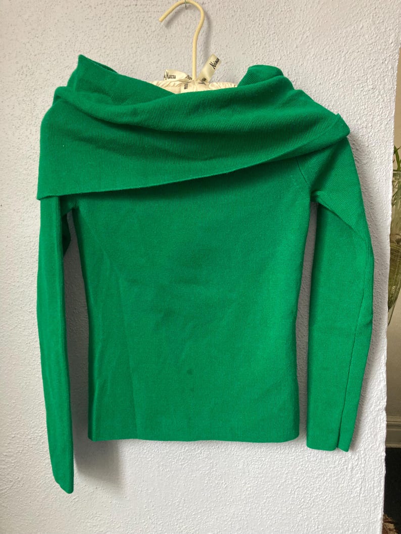 May include: A vibrant green off-the-shoulder jumper with long sleeves. The garment is suspended on a beige hanger against a light grey wall. The neckline is wide and folded over, creating a cowl neck effect.