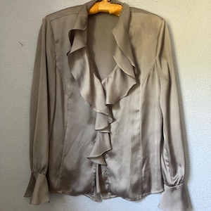 May include: A long-sleeved, taupe-colored blouse with a ruffled front detail. The blouse has a satin-like sheen, a collared neckline, and flared cuffs. It hangs on a vintage yellow hanger against a neutral wall.
