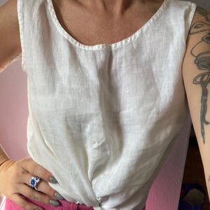 May include: A white linen tank top with a scoop neckline. The top has a twisted knot detail at the waist. The model is wearing pink pants and a blue and white ring.