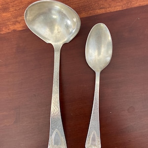May include: A silver ladle and spoon set. The ladle has a large, oval bowl and a long handle with a textured pattern. The spoon has a similar handle design and a smaller, oval bowl. Both utensils have decorative detailing at the base of the handle.
