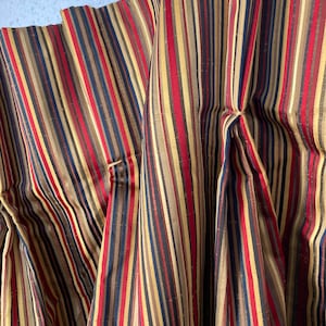 May include: Close-up of a fabric with vertical stripes in red, navy, gold, and brown. The fabric is pleated, creating folds and shadows. The stripes are of varying widths, adding visual interest to the textile.