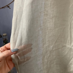 May include: Close-up of a sheer, light beige fabric. The fabric has a textured weave and appears to be linen or a similar material. The fabric is held up, allowing light to pass through, highlighting its transparency.