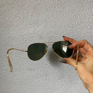 Op de afbeelding: Goudkleurige pilotenbril met donkergroene glazen. De zonnebril heeft een klassieke druppelvorm en een dun, goudkleurig metalen montuur. De glazen zijn donkergroen van kleur.