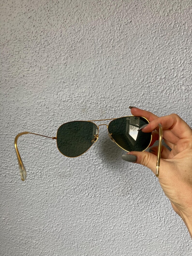 Op de afbeelding: Goudkleurige pilotenbril met donkergroene glazen. De zonnebril heeft een klassieke druppelvorm en een dun, goudkleurig metalen montuur. De glazen zijn donkergroen van kleur.