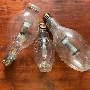 May include: Three clear glass light bulbs with metal bases are arranged on a wooden surface. The bulbs vary in size and shape, with visible internal filaments and support structures. The bases are gold and silver.