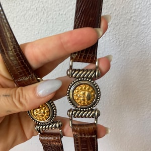 May include: A brown textured leather belt featuring two circular gold-toned sun face medallions. Each medallion is framed by a silver-toned twisted rope border and connected to the leather strap by silver-toned metal hardware links.