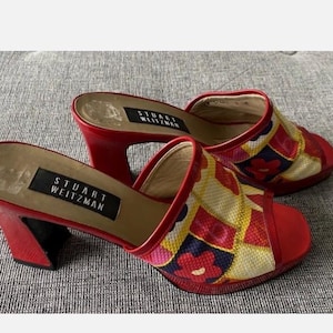 May include: A pair of red Stuart Weitzman heeled mules. The open-toe shoes feature a colorful floral pattern on the upper and a block heel. The brand name is visible on the insole.