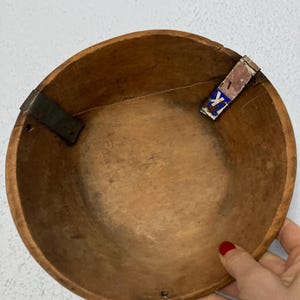 May include: A round, wooden bowl with a light brown finish. The bowl has a simple design with metal brackets on the sides. A small piece of paper with blue and white text is attached to the rim. The bowl is held by a person.