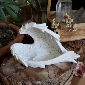 May include: White ceramic angel wing sculpture with detailed feather texture. The wings are open and form a hollow space in the centre. The sculpture is sitting on a wooden surface.