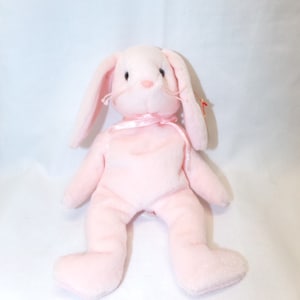 May include: Pink plush bunny rabbit with long floppy ears. The bunny has a pink ribbon around its neck and a red and white tag attached to its ear.