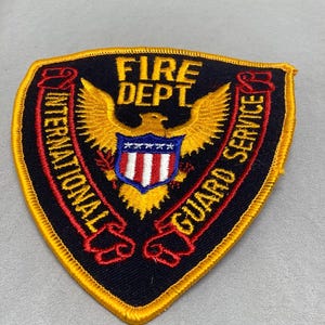 Può includere: Una toppa ricamata nera e gialla con le parole "Fire Dept" e "International Guard Service" in un design a banner curvo. La toppa presenta un'aquila dorata con uno scudo al centro.