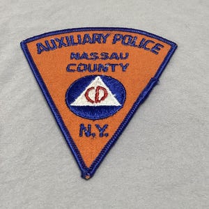 May include: Orange and blue embroidered patch with the text "Auxiliary Police Nassau County NY" and a blue circle with a white triangle and red letter "D" inside.
