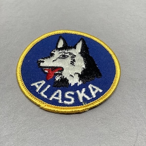 May include: A blue and yellow embroidered patch with a white and black husky dog head and the word "ALASKA" in white lettering.