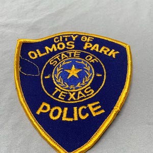 May include: A blue and gold embroidered patch with the text "City of Olmos Park, State of Texas, Police".