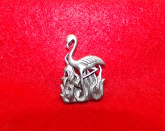 May include: A silver-toned metal brooch featuring a detailed flamingo standing in a bed of stylized reeds.