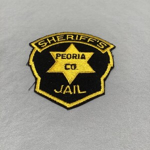 May include: A black and gold Sheriff's patch with the text "SHERIFF'S", "PEORIA CO", and "JAIL". The patch features a gold star in the center.