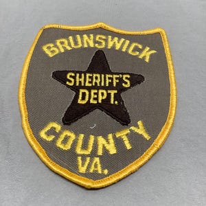 Può includere: Una toppa ricamata marrone e gialla con il testo "Brunswick Sheriff's Dept. County VA."