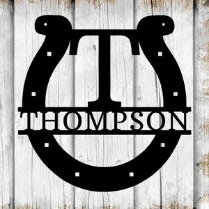 May include: Black metal horseshoe with the name "THOMPSON" and the letter "T" in the center. The design is set against a white and brown wood-grain background. The horseshoe has square cutouts.