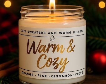 May include: A lit, glass jar candle with a white label featuring gold text that reads "Warm & Cozy." The label also includes the phrases "Cozy Sweaters and Warm Hearts" and scent notes: orange, pine, cinnamon, and clove. The candle is surrounded by holiday decorations.