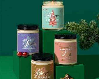 May include: Four glass jar candles with black lids, displayed on green blocks. One candle is lit, labelled "Merry Christmas." Other labels read "Let it Snow," "Naughty List," and "Spiced Gingerbread." A gingerbread star and sprig of greenery complete the festive scene.
