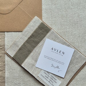 Op de afbeelding: Een close-up van een staal van haverkleurig linnen stof met de tekst "Mira - Oatmeal Oeko-Tex 100% Linen width: 149cm AVLEN Linen Home Living" gedrukt op een wit label. De stof wordt getoond op een beige achtergrond.