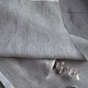 May include: A grey and white striped linen apron with a white trim. The apron is folded in half and has a single strap.