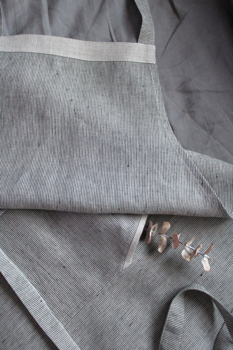 May include: A grey and white striped linen apron with a white trim. The apron is folded in half and has a single strap.
