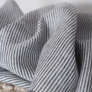 May include: A piece of fabric featuring a pattern of thin, vertical black and white stripes. The material is draped in soft, flowing folds, highlighting the consistent linear design and the texture of the woven textile across the surface.