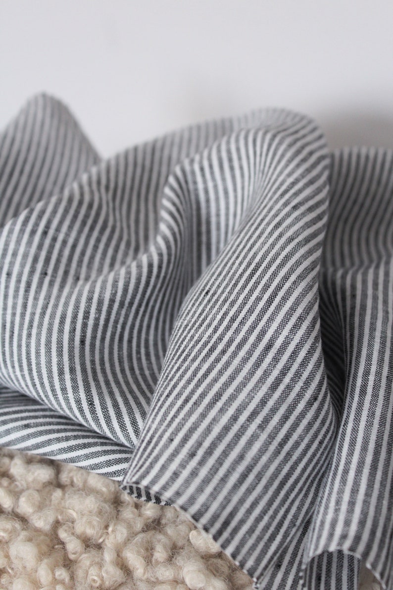 May include: A piece of fabric featuring a pattern of thin, vertical black and white stripes. The material is draped in soft, flowing folds, highlighting the consistent linear design and the texture of the woven textile across the surface.