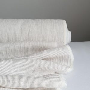 May include: A roll of off-white fabric featuring a subtle, fine vertical pinstripe pattern in a light gray tone. The textile is tightly wound on a cylindrical core, with several layers of the material draped forward, showcasing a soft texture.
