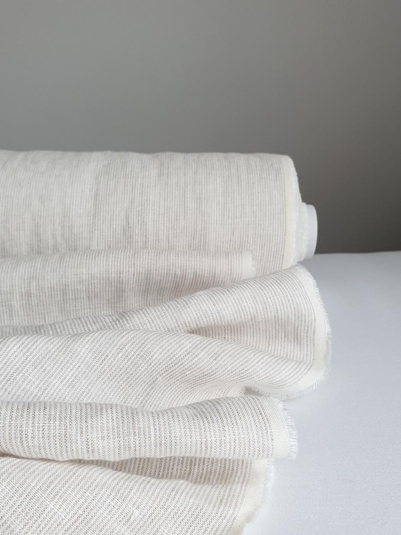 May include: A roll of off-white fabric featuring a subtle, fine vertical pinstripe pattern in a light gray tone. The textile is tightly wound on a cylindrical core, with several layers of the material draped forward, showcasing a soft texture.