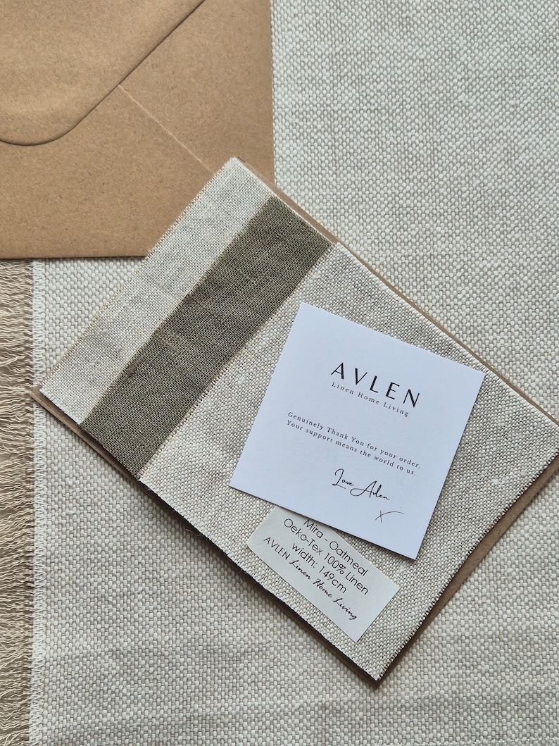 Op de afbeelding: Een close-up van een staal van haverkleurig linnen stof met de tekst "Mira - Oatmeal Oeko-Tex 100% Linen width: 149cm AVLEN Linen Home Living" gedrukt op een wit label. De stof wordt getoond op een beige achtergrond.