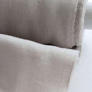 May include: A roll of light beige fabric with a visible woven texture and frayed edges. The material is tightly wound into a cylindrical shape, with a portion of the fabric unrolled to display the weave and the soft, neutral color of the textile.
