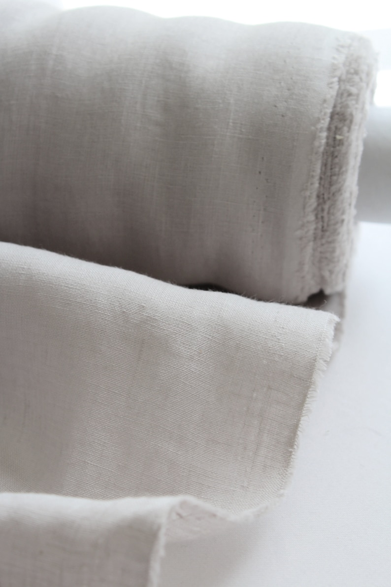 May include: A roll of light beige fabric with a visible woven texture and frayed edges. The material is tightly wound into a cylindrical shape, with a portion of the fabric unrolled to display the weave and the soft, neutral color of the textile.